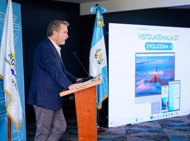 Director del Inguat, Harris Whitbeck, presenta en conferencia de prensa la renovación del portal oficial de turismo visitguatemala.gt, con inteligencia artificial para crear itinerarios personalizados, frente a la bandera de Guatemala.