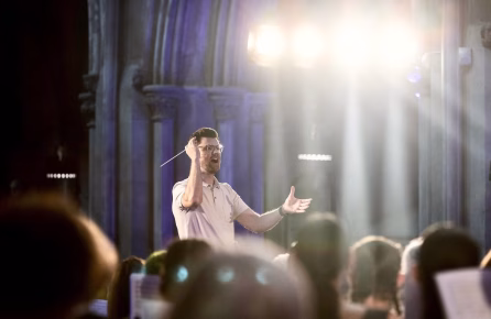 Jake Runestad dirige con batuta en mano durante el concierto YOUR SOUL IS SONG en Guatemala, rodeado de coristas, bajo haces de luz dramáticos en un recinto histórico