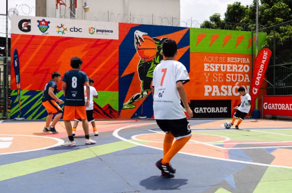 Jóvenes juegan fútbol en una cancha comunitaria decorada con un mural de Estadios del Barrio en La Palmita, zona 5